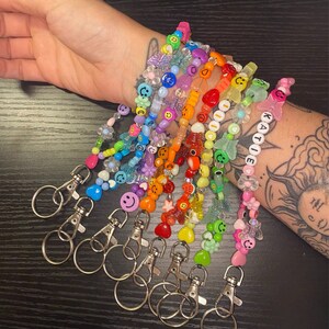 May include: A collection of colorful beaded keychains with various charms, including smiley faces, flowers, and butterflies. One keychain has the name "KATIE" spelled out in white beads. Each keychain has a silver clasp.