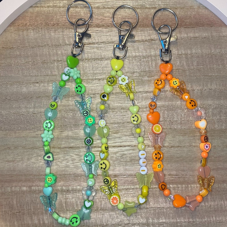May include: Three beaded keychains with silver clasps. The keychains feature colorful beads in shades of green, yellow, and orange. The beads are shaped like smiley faces, butterflies, flowers, and hearts.