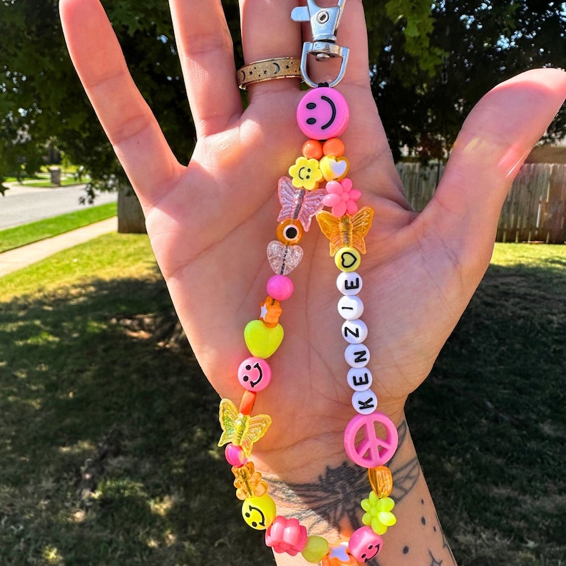 May include: A beaded keychain with a silver clasp. The keychain features a variety of colorful beads, including smiley faces, hearts, flowers, and butterflies. The name "KENZIE" is spelled out with white letter beads. The color scheme includes pink, orange, yellow, and clear beads.