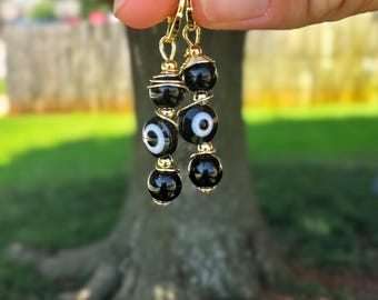 May include: Gold-toned hoop earrings with black and white evil eye beads. The earrings feature a combination of glossy black beads and beads with a white eye design, strung together with gold wire.