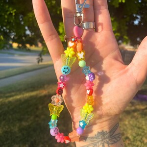 May include: A colorful beaded keychain with a silver clasp. The keychain features a variety of beads, including smiley faces, butterflies, stars, flowers, and hearts in a rainbow of colors. The beads are strung together to create a fun and playful accessory.