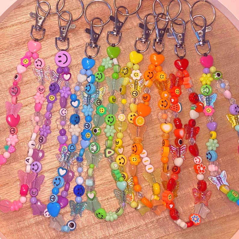 May include: A collection of beaded keychains in a rainbow of colors. Each keychain features a silver clasp and is adorned with various beads, including smiley faces, butterflies, hearts, and flowers. The keychains are arranged on a wooden surface.