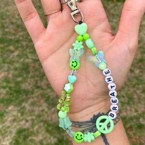 May include: A green beaded keychain with a silver clasp. The keychain features various green beads, including smiley faces, butterflies, hearts, and flowers. The word "breathe" is spelled out in white letter beads, and a peace sign is also included.