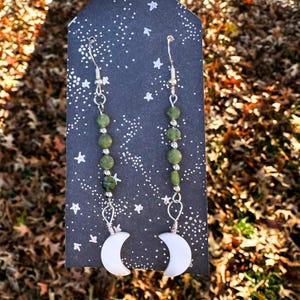 May include: A pair of silver earrings with green gemstone beads and white crescent moon charms.