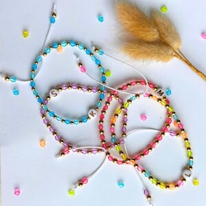 Op de afbeelding: Vijf kleurrijke kralenarmbanden met gouden accenten en letterbedels. De armbanden zijn in tinten blauw, roze, paars, geel en groen. De bedels spellen de letters S, K, M en L.