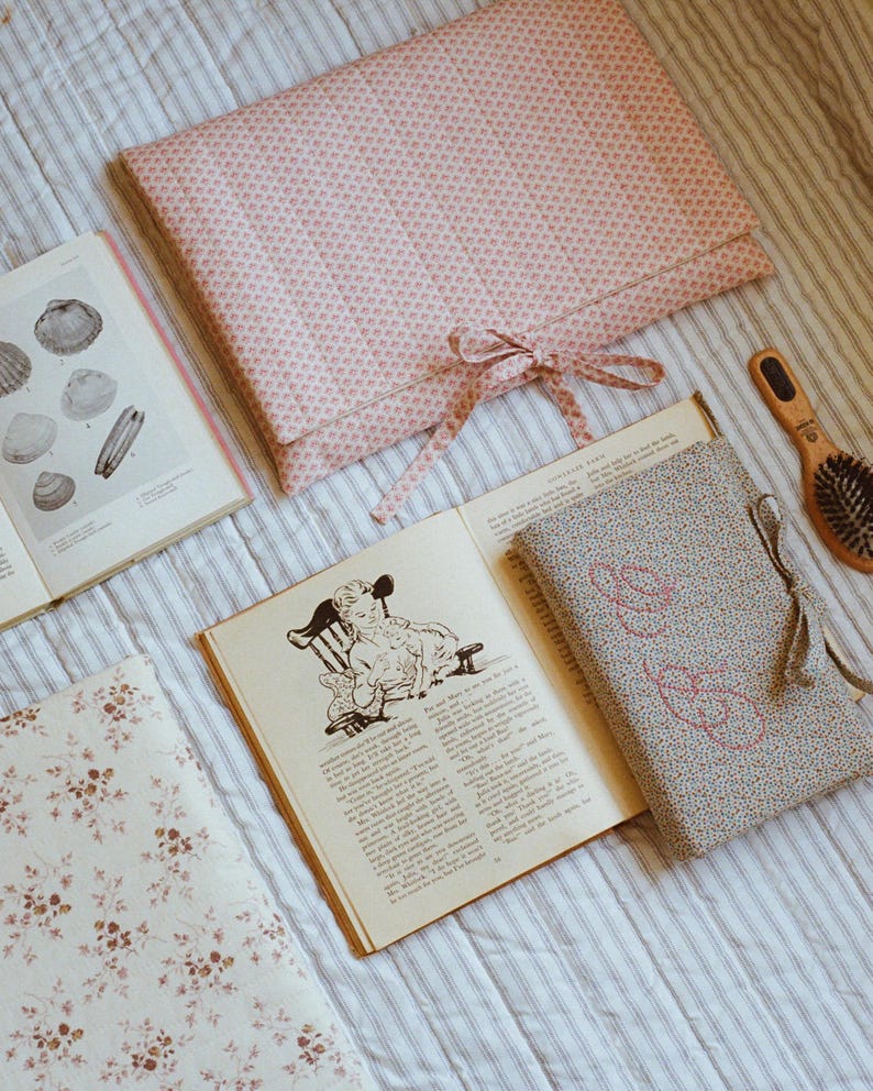 K&ouml;nnte beinhalten: Eine Sammlung von Vintage-Artikeln, darunter offene B&uuml;cher mit Illustrationen und Text, ein rosa Stoff mit Blumenmuster und eine Holzb&uuml;rste. Ein gro&szlig;er rosa Stoffumschlag mit Schleife ist ebenfalls vorhanden.