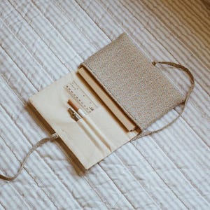 K&ouml;nnte beinhalten: Ge&ouml;ffnete Federtasche mit floralem Muster und beigem Innenraum, mit Lineal, Bleistiften und einem Stift. Die Tasche ist offen und zeigt ein Notizbuch. Die Tasche hat einen passenden Riemen und liegt auf einer gestreiften, gesteppten Oberfl&auml;che.