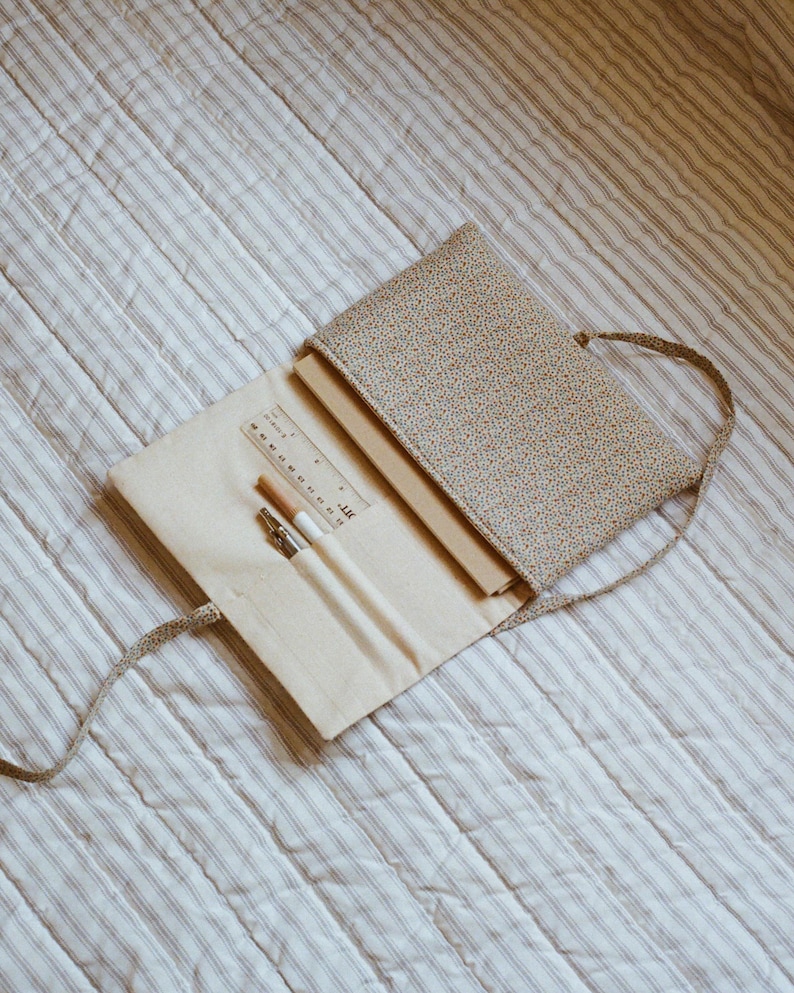 K&ouml;nnte beinhalten: Ge&ouml;ffnete Federtasche mit floralem Muster und beigem Innenraum, mit Lineal, Bleistiften und einem Stift. Die Tasche ist offen und zeigt ein Notizbuch. Die Tasche hat einen passenden Riemen und liegt auf einer gestreiften, gesteppten Oberfl&auml;che.