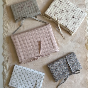 K&ouml;nnte beinhalten: F&uuml;nf gesteppte Stoffh&uuml;llen in verschiedenen floralen und gemusterten Designs. Die H&uuml;llen sind in Rosa-, Wei&szlig;- und Blaut&ouml;nen gehalten und haben Schleifenb&auml;nder. Eine H&uuml;lle hat gestickte Initialen.