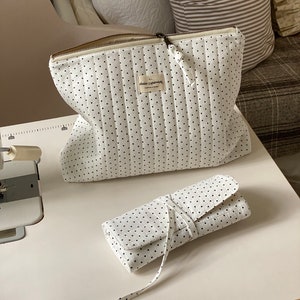 Peut inclure: Deux sacs matelassés blancs à pois noirs. Le plus grand sac a une fermeture éclair et une étiquette qui indique "nolie cippewa". Le plus petit sac est roulé et attaché avec un ruban blanc.