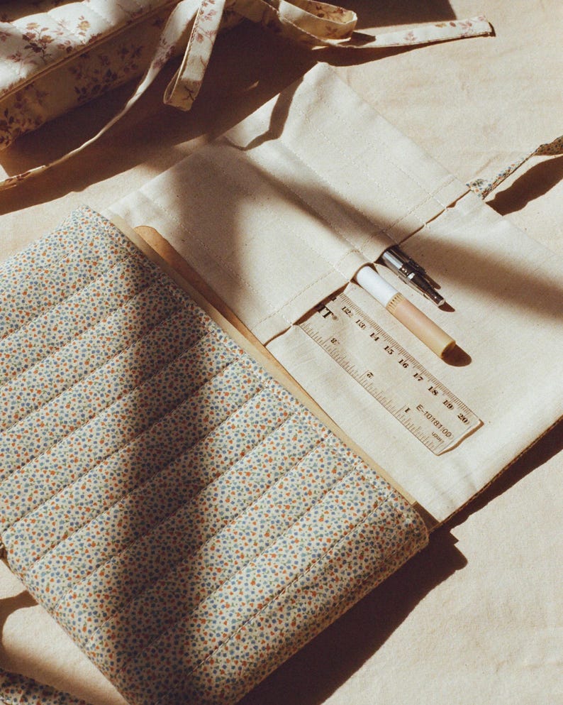 K&ouml;nnte beinhalten: Cremefarbene Federtasche aus Canvas mit floral gemusterter Stepph&uuml;lle. Ein Lineal, Stift und Bleistift sind sichtbar. Die Tasche ist ge&ouml;ffnet und zeigt ihren Inhalt. Das Design ist weich und vintage.