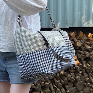 Peut inclure: Sac à bandoulière matelassé à motif vichy noir et blanc. Le sac est doté d'un rabat et d'une bandoulière assortie. La personne porte une chemise blanche et un short en jean.