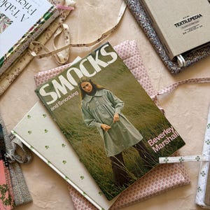 K&ouml;nnte beinhalten: Eine Sammlung von B&uuml;chern und Buchumschl&auml;gen mit floralen und gemusterten Designs. Die Buchumschl&auml;ge sind in verschiedenen Farben gehalten, darunter Rosa, Gr&uuml;n und Beige. Ein Buch tr&auml;gt den Titel "Smocks and Smocking" und ein anderes "A Table for Friends".