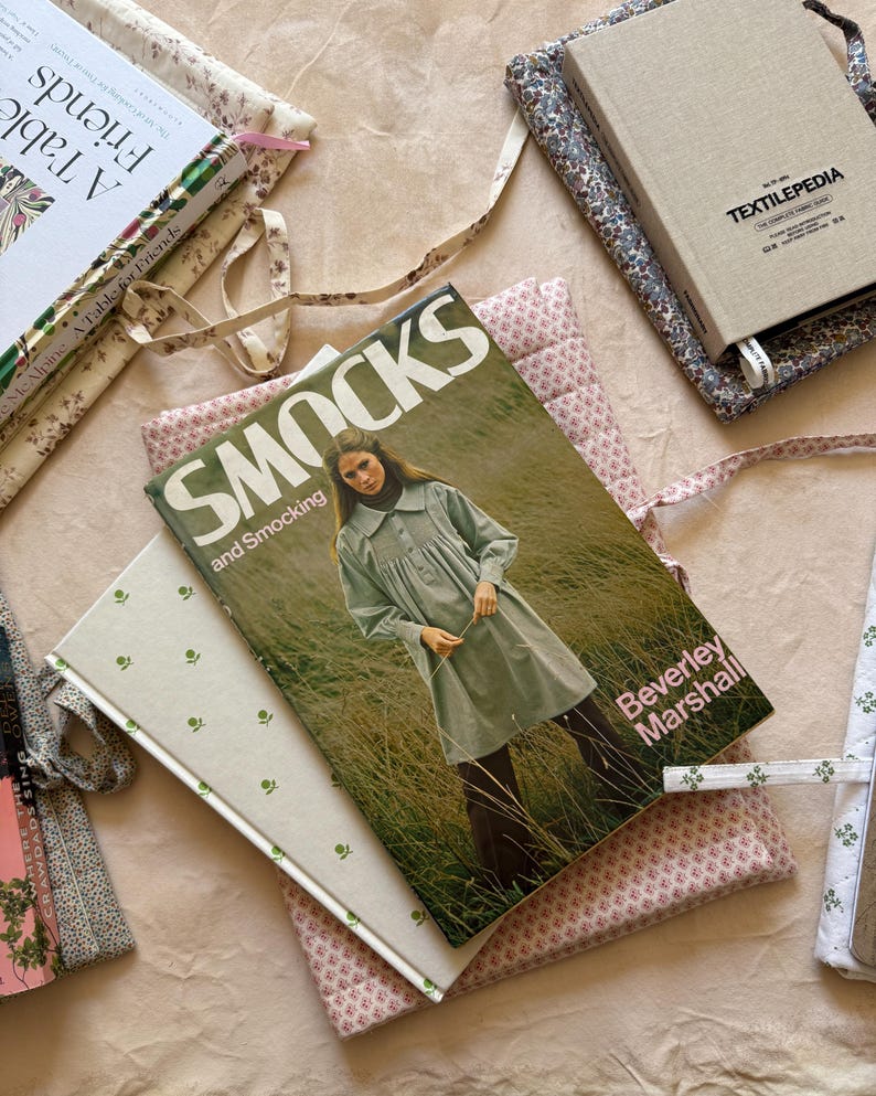 K&ouml;nnte beinhalten: Eine Sammlung von B&uuml;chern und Buchumschl&auml;gen mit floralen und gemusterten Designs. Die Buchumschl&auml;ge sind in verschiedenen Farben gehalten, darunter Rosa, Gr&uuml;n und Beige. Ein Buch tr&auml;gt den Titel "Smocks and Smocking" und ein anderes "A Table for Friends".