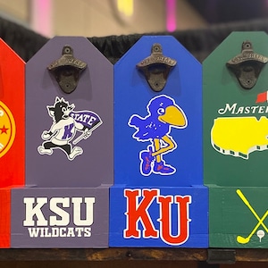 May include: A row of eight wooden wall-mounted bottle openers, each featuring a unique sports or themed graphic. The designs include team logos, mascots, and symbols in various colors like blue, red, black, green, and teal, all with metal openers.