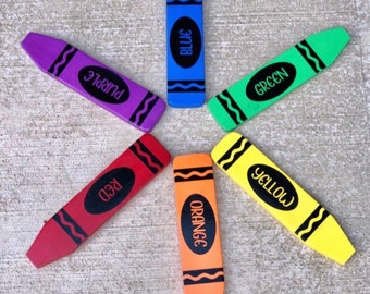 May include: Six crayon-shaped objects arranged in a star pattern. Each crayon is a different color: blue, green, yellow, orange, red, and purple. Each features a black oval in the center with the color name printed in black text and black wavy lines.