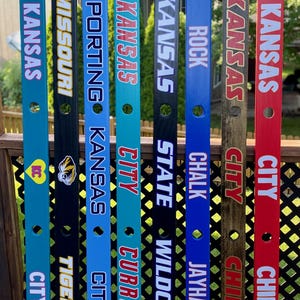 May include: A collection of eight decorative wooden signs, each painted in vibrant colors and featuring team names and logos. The signs are approximately 48 inches tall and are designed with a ski-like shape, perfect for displaying team pride.