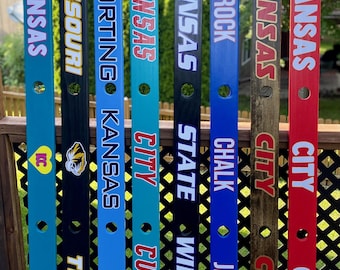 Könnte beinhalten: Acht vertikale Holzschilder mit Sport-Teamnamen und Logos. Die Schilder sind in verschiedenen Farben wie Türkis, Schwarz, Blau und Rot lackiert. Jedes Schild zeigt Texte wie Kansas, Missouri, Sporting, City, State, Wildcats, Rock, Chalk, Jayhawk und Chiefs.