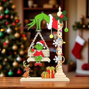 Puede incluir: Adorno navideño de madera con el Grinch con gorro de Papá Noel, sosteniendo una taza, sentado en un columpio. La decoración incluye una mano, luces, copos de nieve y un perro pequeño. Los regalos están en la base, con un árbol de Navidad al fondo.
