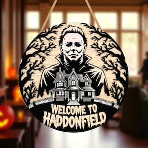 Könnte beinhalten: Rundes Halloween-Holzschild mit einem Schwarz-Weiß-Bild von Michael Myers, einem Haus und dem Text "WELCOME TO HADDONFIELD". Das Schild wird an einem Naturseil aufgehängt. Der Hintergrund ist verschwommen, mit einem Kürbis und einer Person.