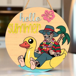 Peut inclure: Panneau rond en bois avec l'inscription "hello summer" en bleu et jaune. Un personnage de dessin animé avec un chapeau et des lunettes de soleil flotte sur un canard gonflable jaune. Une fleur d'hibiscus rose et un palmier sont également présents.