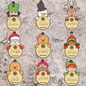 Puede incluir: Nueve adornos navideños personalizados con varios personajes, incluyendo un hombre de jengibre, un muñeco de nieve, un reno, Papá Noel y un elfo. Cada adorno tiene un diseño de copo de nieve y un nombre en un círculo de madera.