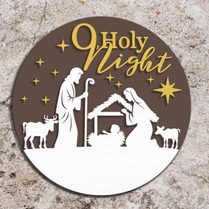 May include: A round, layered wooden sign with a Christmas nativity scene. The sign features the words "O Holy Night" in gold lettering, with a brown background and white cut-out figures of Mary, Joseph, baby Jesus, and animals.