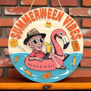 May include: Round wooden sign with the text "SUMMERWEEN VIBES" in orange. The sign features a cartoon character in a red shirt and sunglasses, floating on a pink flamingo floatie, holding a drink. Candy and a "BOO" treat bag are also depicted.