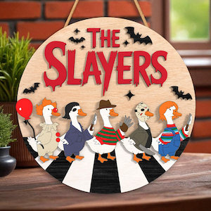 May include: A round wooden sign with the text "THE SLAYERS" in red, featuring cartoon ducks dressed as horror movie characters. The sign includes bats and stars, with a rope for hanging. The ducks are on a black and white striped path.