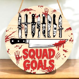 May include: Round wooden sign with a light wood grain background. The sign features a collection of black and white knives and a large butcher knife with a blood-red blade. The words "SQUAD GOALS" are in red. A rope is attached for hanging.