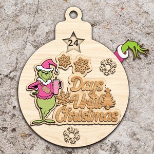 Può includere: Ornamento natalizio in legno con un personaggio verde in abito rosa che tiene un bicchiere rosa. L'ornamento ha un ritaglio a forma di stella con il numero 24, fiocchi di neve e la scritta "Days Until Christmas". Una mano verde con un polsino bianco è a destra.