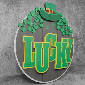 May include: A round wooden sign with a gray background and the word "LUCKY" in green letters with a yellow outline. The sign is decorated with green shamrocks and a green leprechaun hat with a gold buckle.