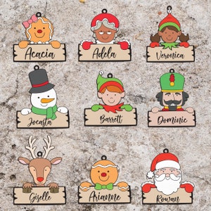 Op de afbeelding: Negen kerstversieringen met verschillende figuren die borden met namen vasthouden. De figuren omvatten een peperkoekpersoon, een vrouw met een rode hoed, een elf, een sneeuwpop, een notenkraker, een rendier, de kerstman en twee andere peperkoekpersonen. De namen op de borden zijn Acacia, Adela, Veronica, Jocasta, Barrett, Dominic, Giselle, Arianne en Rowan.