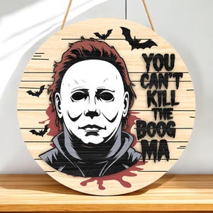 May include: Round wooden Halloween sign featuring a graphic of a masked character with red hair and the text "YOU CAN'T KILL THE BOOG MA". Includes bat silhouettes and a blood splatter design.