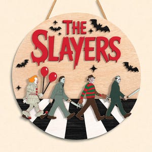 May include: Round wooden sign with the text "THE SLAYERS" in red, featuring horror movie characters walking on a black and white crosswalk. Includes bats and stars.