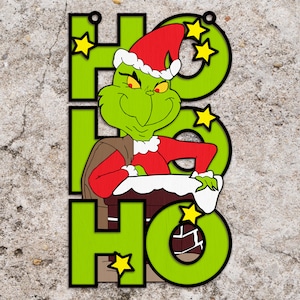 Puede incluir: Adorno navideño verde y rojo con el Grinch de la película "El Grinch". El adorno tiene forma de árbol de Navidad y dice "HO HO" en él.