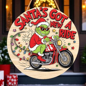May include: Round wooden sign with the text "SANTA'S GOT A NEW RIDE". The sign features a cartoon Grinch character in a Santa suit riding a red motorcycle, with Christmas lights trailing behind.