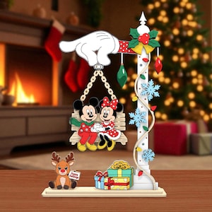 May include: A Christmas-themed decoration featuring Mickey and Minnie Mouse on a swing, held by a white gloved hand. The decoration includes a reindeer holding a sign that says "Happy Holidays", gifts, and a decorative pole with snowflakes and ornaments.