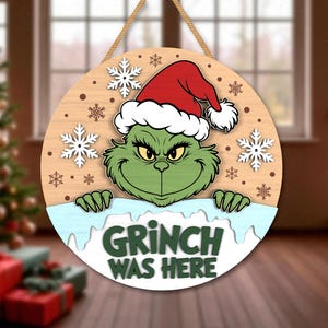 May include: Round wooden Christmas sign featuring the Grinch wearing a Santa hat. The Grinch is green with a mischievous expression. The sign reads "Grinch Was Here" in green letters, with snowflakes and a light blue border.