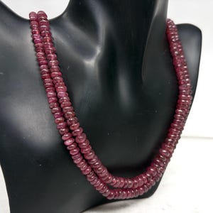 May include: A necklace of deep red, polished gemstones. The necklace is composed of multiple strands of small, round beads. The beads are a rich, dark red color, and the necklace is displayed on a black mannequin.