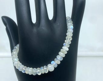May include: A delicate bracelet composed of small, round, iridescent moonstone beads. The bracelet is displayed on a black hand-shaped display, showcasing the subtle colors and textures of the gemstones. The background is a soft white.