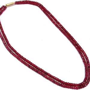 Puede incluir: Un collar de tres hilos compuesto por pequeñas cuentas facetadas de color rojo intenso. El collar se asegura con un cierre dorado. Las cuentas tienen un tamaño y forma uniformes, creando una apariencia uniforme. El collar se muestra sobre un fondo blanco.