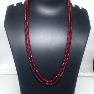 May include: A double-strand necklace composed of small, faceted, deep red beads. The necklace is displayed on a black mannequin, showcasing its length and drape. The beads catch the light, creating a sparkling effect.