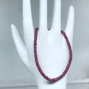 Peut inclure: Un bracelet délicat composé de petites pierres précieuses roses foncées et facettées. Le bracelet est présenté sur un présentoir blanc en forme de main, mettant en valeur la couleur et le design du bijou. Les pierres semblent être de taille uniforme et enfilées.