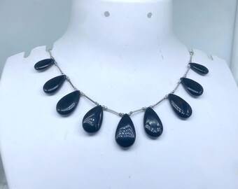 Puede incluir: Un collar con cuentas negras pulidas en forma de lágrima ensartadas en una cadena plateada. Las cuentas están espaciadas uniformemente y tienen un acabado brillante, creando un diseño simple pero elegante. El collar se exhibe en un maniquí blanco.