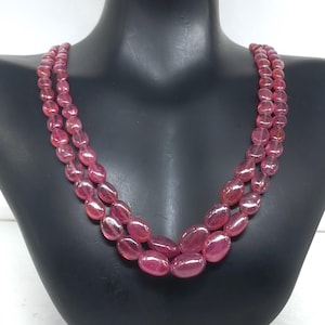 Puede incluir: Un collar de dos hilos con piedras preciosas ovaladas de color rosa rojizo. Las cuentas están ensartadas, creando un efecto en capas. El collar se exhibe en un busto de maniquí negro, mostrando el diseño y el color de la joya.
