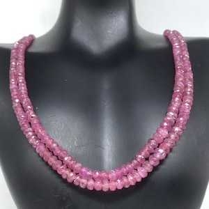 Puede incluir: Un collar de tres hilos compuesto por pequeñas piedras preciosas rosas facetadas. El collar se exhibe sobre un busto de maniquí negro. Las piedras preciosas tienen un aspecto brillante y el color general es un rosa vibrante.