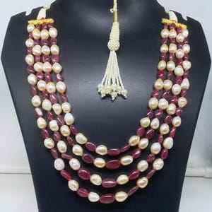 Può includere: Collana a più fili con fili alternati di perle bianche cremose e pietre preziose rosso intenso. La collana è esposta su un manichino nero, mostrando il suo design a strati e i suoi dettagli intricati. Un nappa di perline pende sul retro.