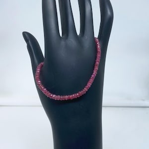 Puede incluir: Una delicada pulsera compuesta por pequeñas cuentas facetadas de color rubí. Las cuentas están ensartadas para formar un hilo flexible. La pulsera se muestra sobre un expositor negro con forma de mano, sobre un fondo blanco.
