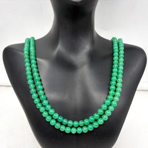 Peut inclure: Un collier à deux rangs composé de perles vertes vives. Les perles sont de taille et de forme uniformes, créant une ligne lisse et continue. Le collier est présenté sur un mannequin noir, mettant en valeur la couleur et le design du bijou.